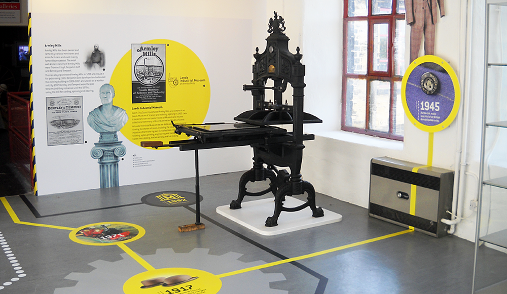 Vintage printing press displayed in a gallery with informational panels and a timeline on the floor.