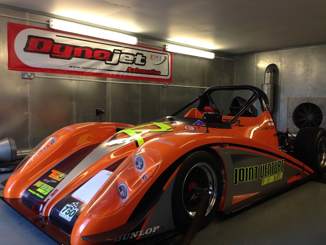 Orange racing car with "Joint Venture" branding inside a garage, featuring a Dynojet banner on the wall.