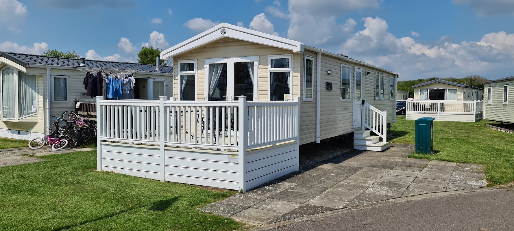 Wooden caravan with a deck, surrounded by green grass and nearby caravans, under a blue sky with fluffy clouds.