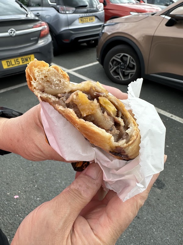 A hand holding a flaky pastry filled with meat and vegetables, with parked cars in the background.