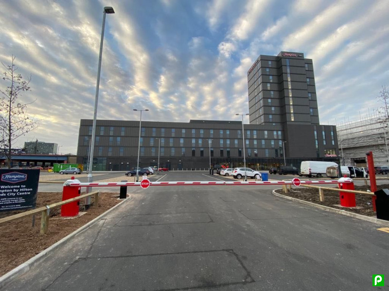 Entrance to JustPark car park with barrier, modern building in background, and dramatic cloud-filled sky.