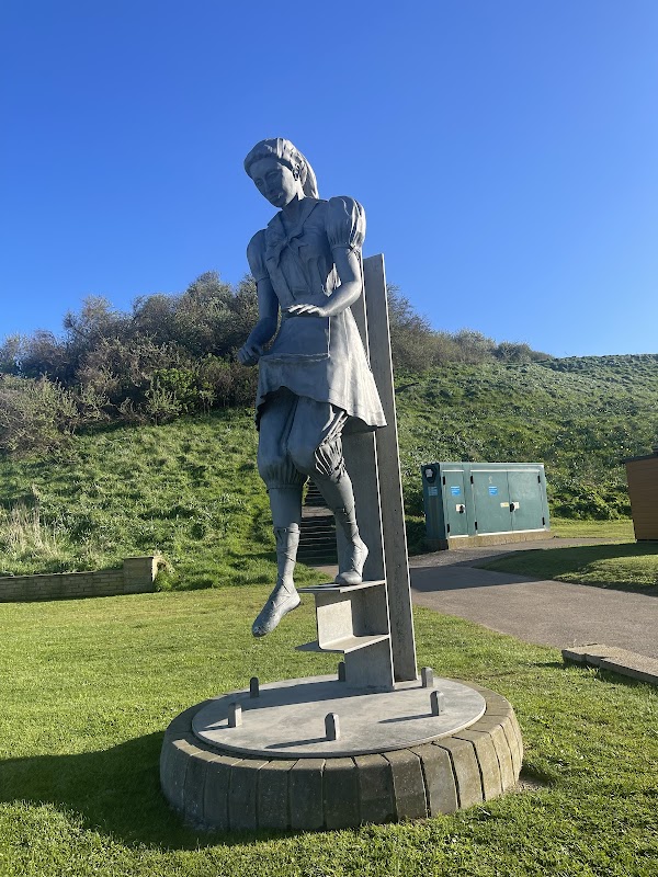 A large statue of a girl in a dress, standing on a pedestal, surrounded by green grass and a clear blue sky.