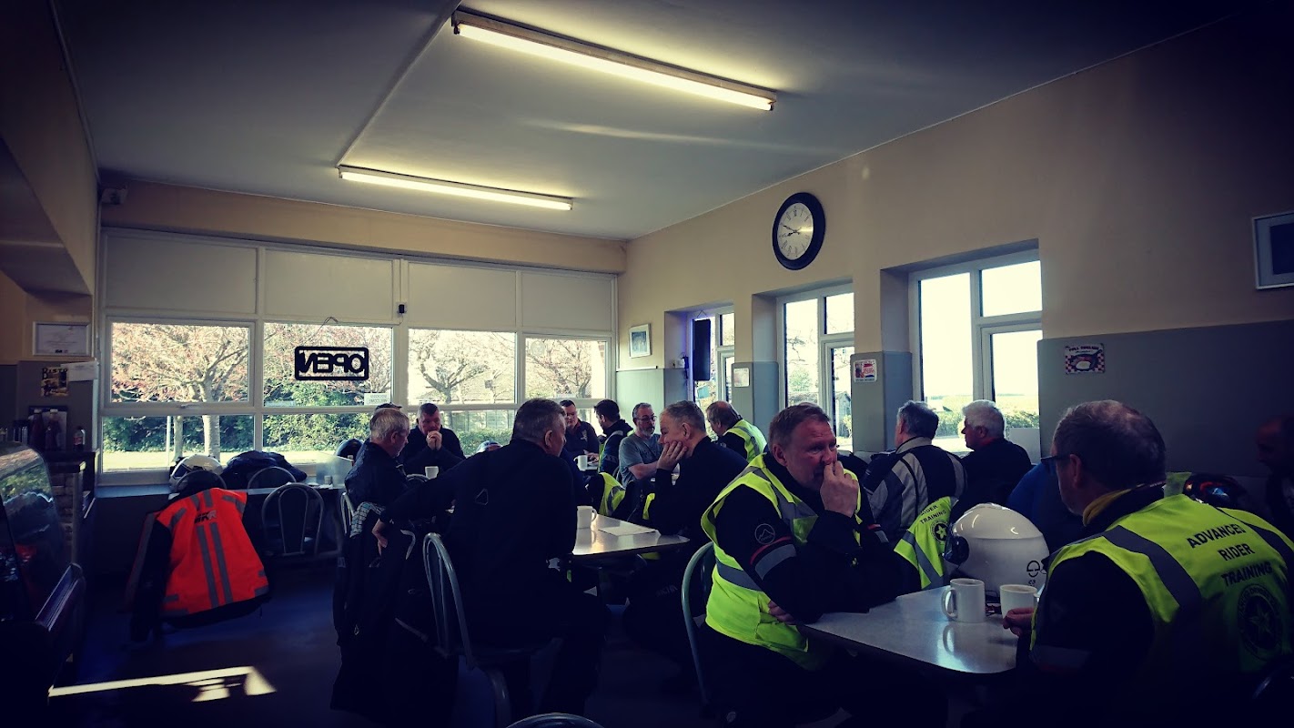 Busy cafe interior with patrons in high-visibility jackets enjoying food and drinks at tables, bright windows letting in light.