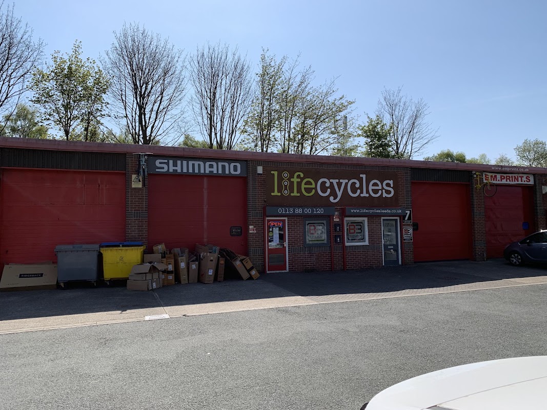 Red storefront with "Shimano" and "lifecycles" signage, surrounded by trees and parked cars. Boxes are stacked outside.