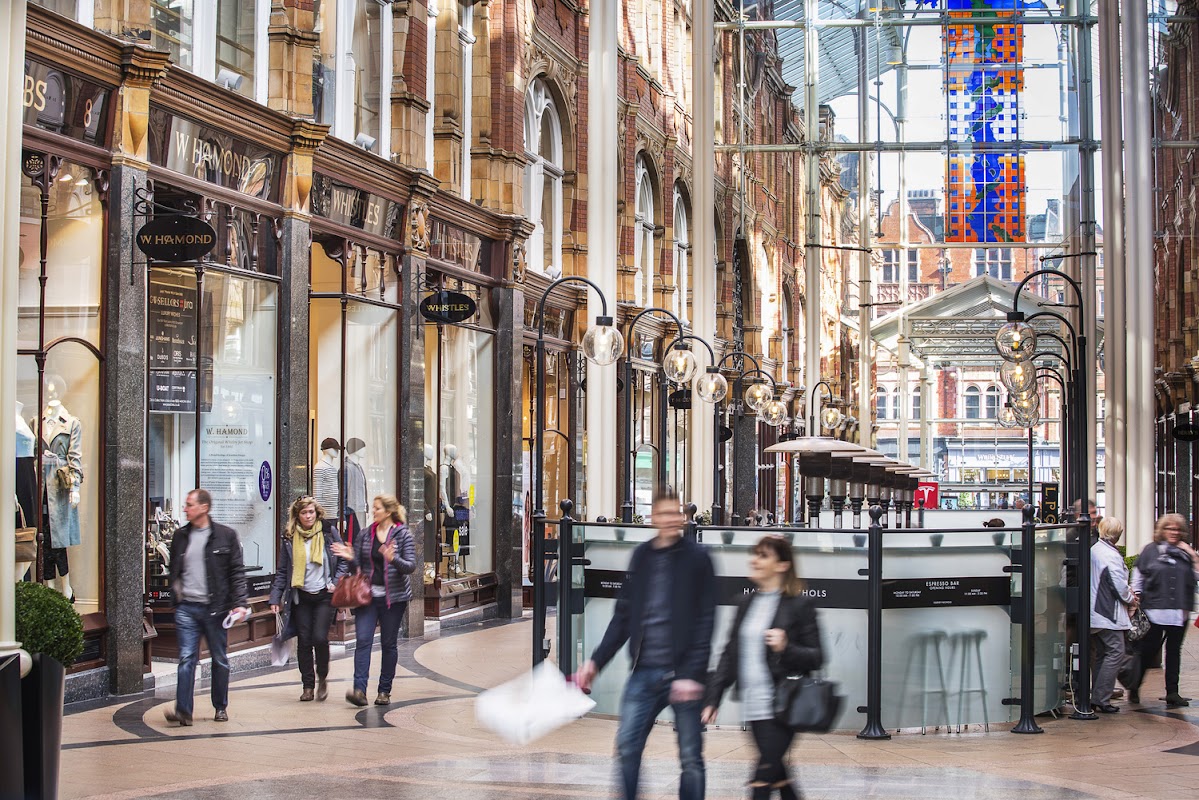 Victorian-style shopping arcade with glass roof, ornate architecture, and shoppers walking through the bustling space.