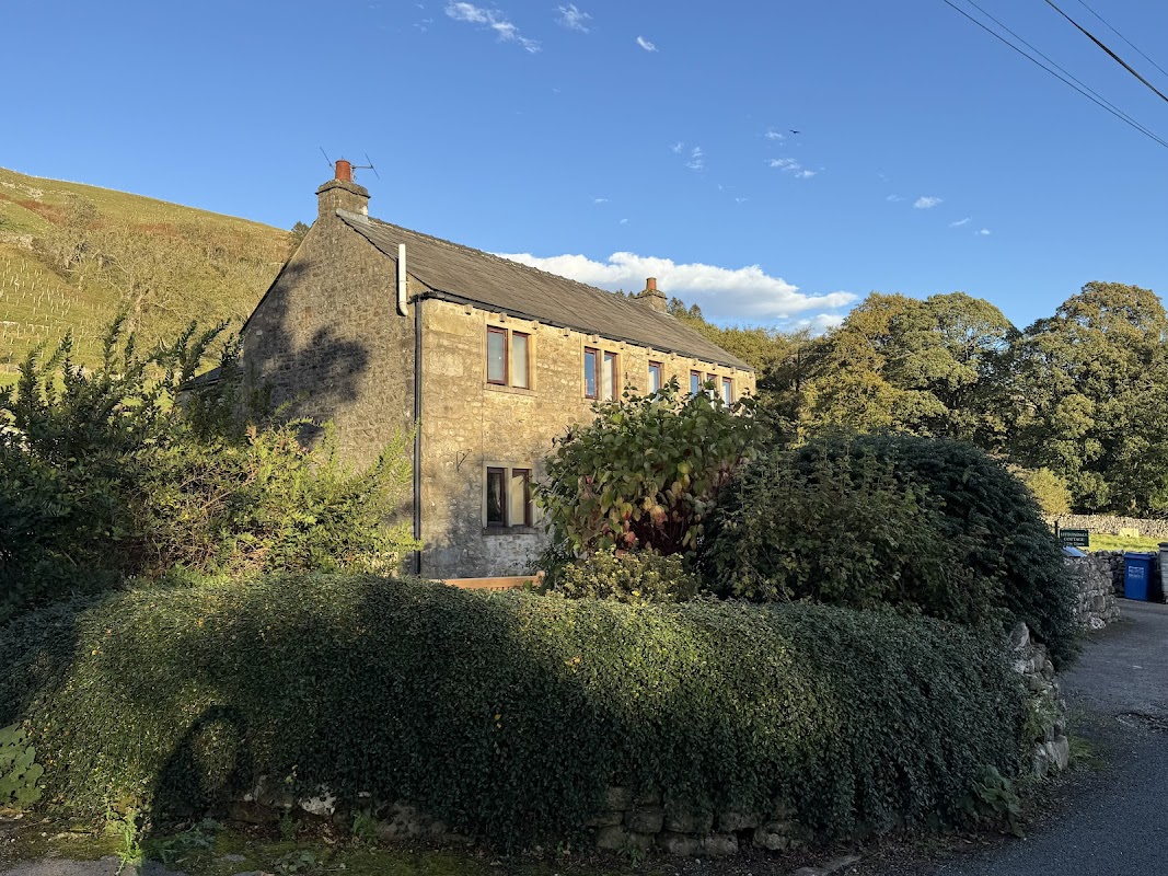 Stone cottage surrounded by greenery, with a clear blue sky and hills in the background, located in Litton, North Yorkshire.