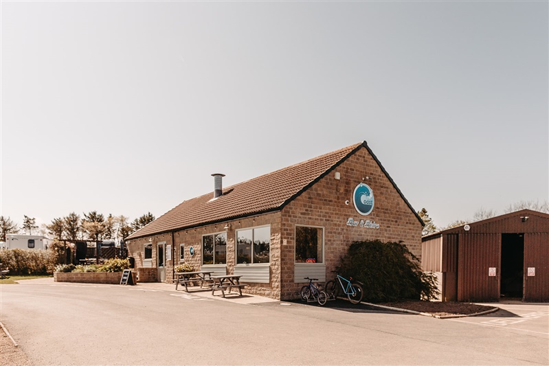 Holiday park building with large windows, outdoor seating, and a bike parked outside in a sunny setting.