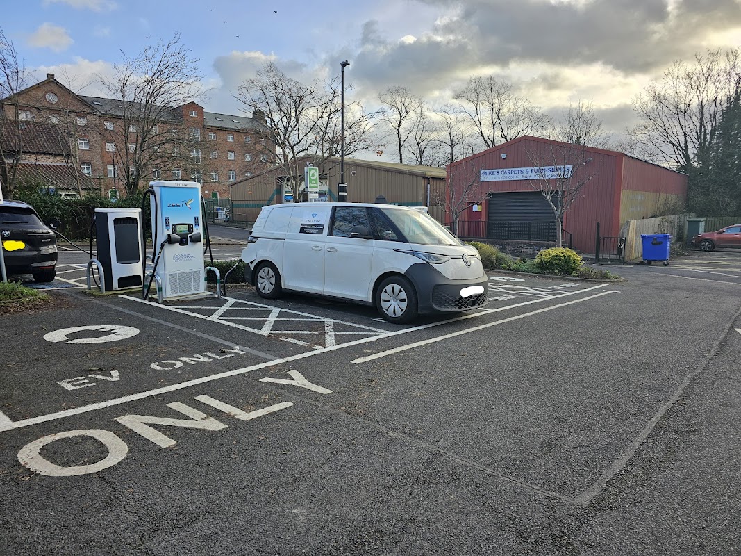 Electric vehicle charging station with a parked van in a car park, surrounded by trees and buildings.