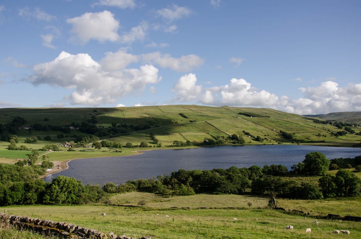 Serene landscape of Semerwater lake surrounded by lush green hills and scattered trees under a blue sky with clouds.