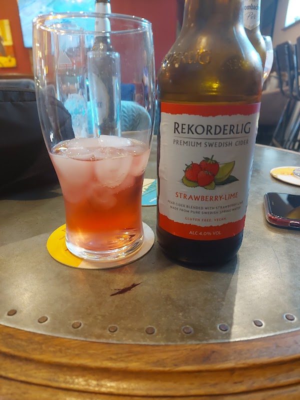 A glass of ice with a pink drink beside a bottle of Rekorderlig Strawberry-Lime cider on a table.