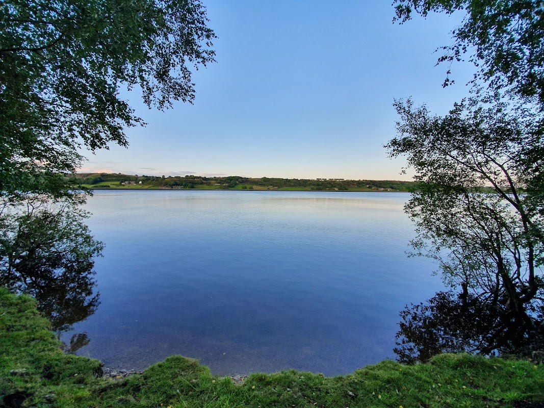 Serene view of Mixenden Reservoir surrounded by trees and a clear blue sky, reflecting calm waters.