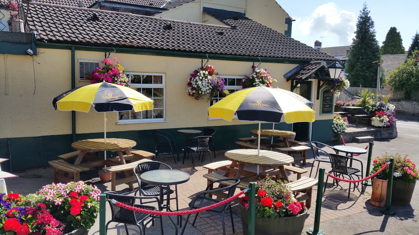 Yellow and black umbrellas shade wooden tables outside the Cross Keys Inn, surrounded by vibrant flower displays.