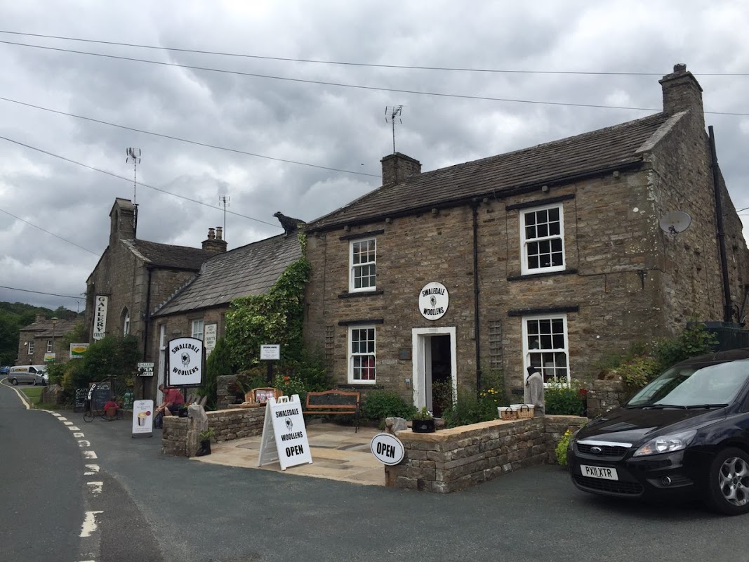 Stone shopfront with "Swaledale Woollens" sign, potted plants, open sign, and parked cars on a cloudy day in Muker.