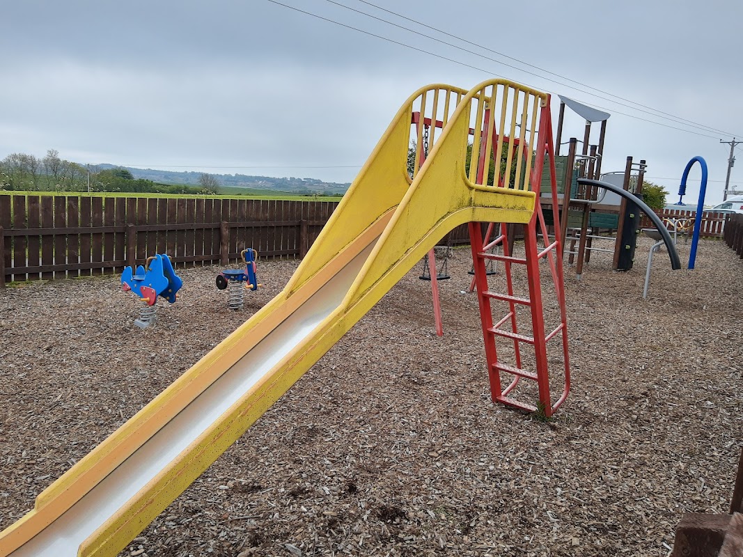 Bright yellow slide and climbing frame in a playground surrounded by wooden fencing and gravel.