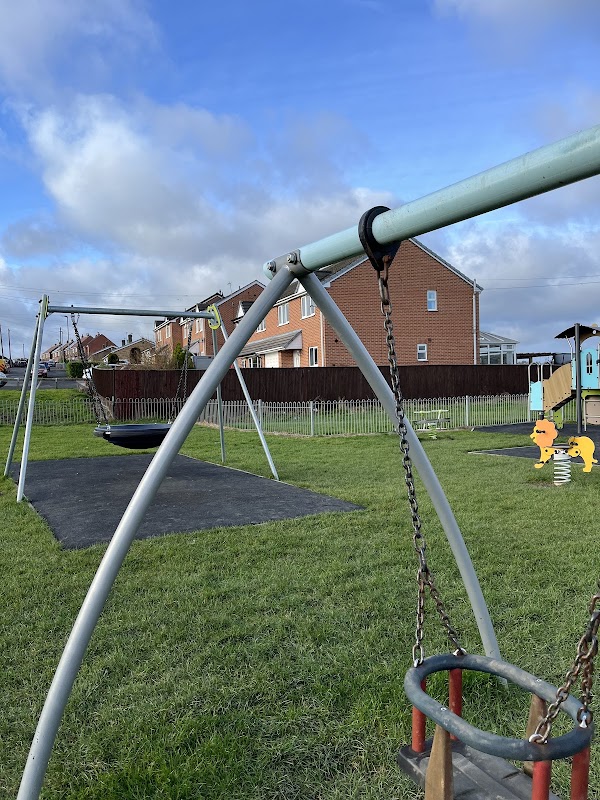 Playground with swings, a spring rider, and grassy areas, surrounded by houses under a blue sky with clouds.