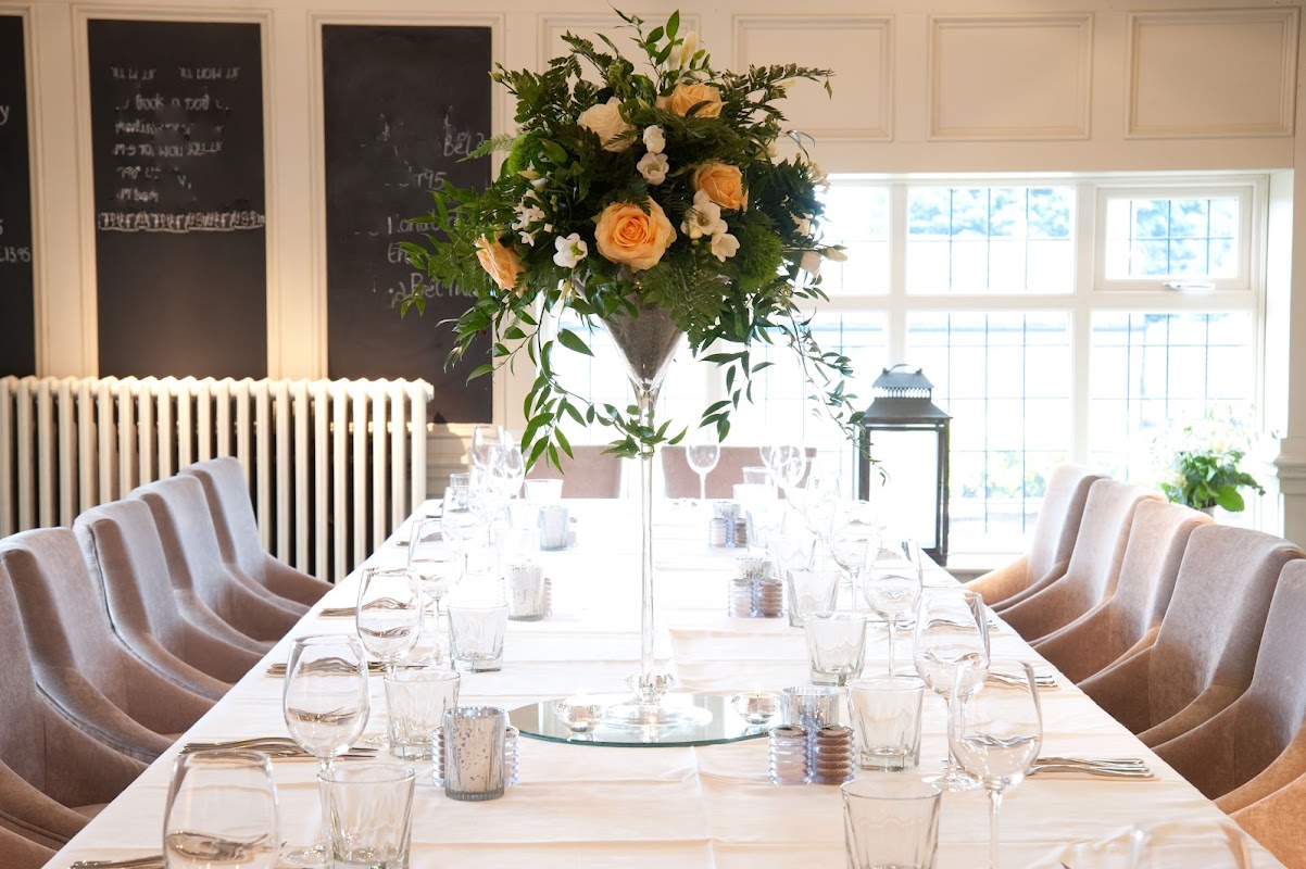 Elegant dining setup with a long table, glassware, and a tall floral centerpiece featuring roses and greenery.