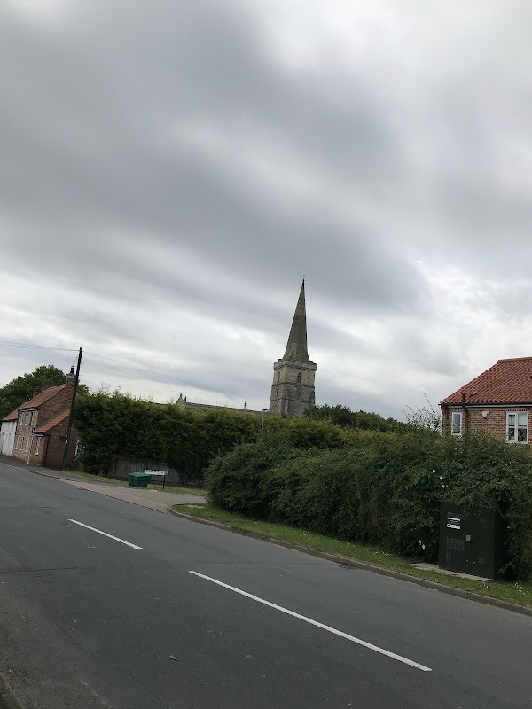Village hall with a church spire in the background, surrounded by greenery and a quiet road in Ottringham, Yorkshire.
