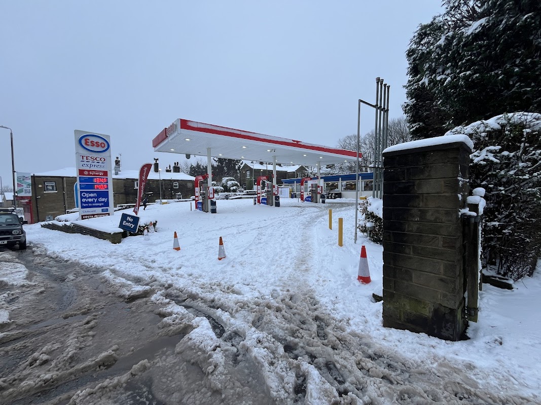 Esso gas station in Ovenden, Yorkshire, surrounded by snow, with visible fuel pumps and a sign indicating open hours.