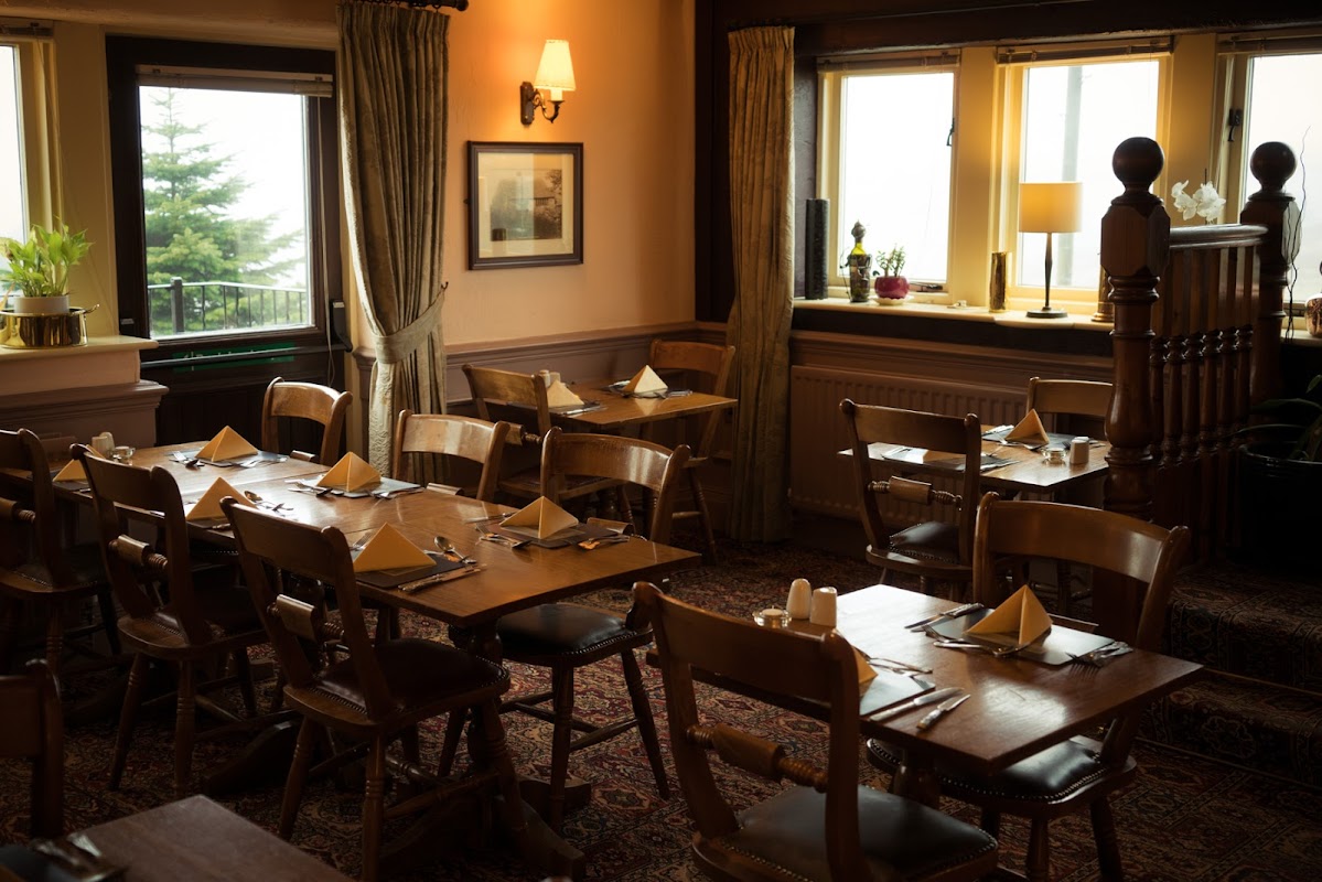 Cozy dining area with wooden tables, chairs, and neatly set napkins, surrounded by warm lighting and large windows.