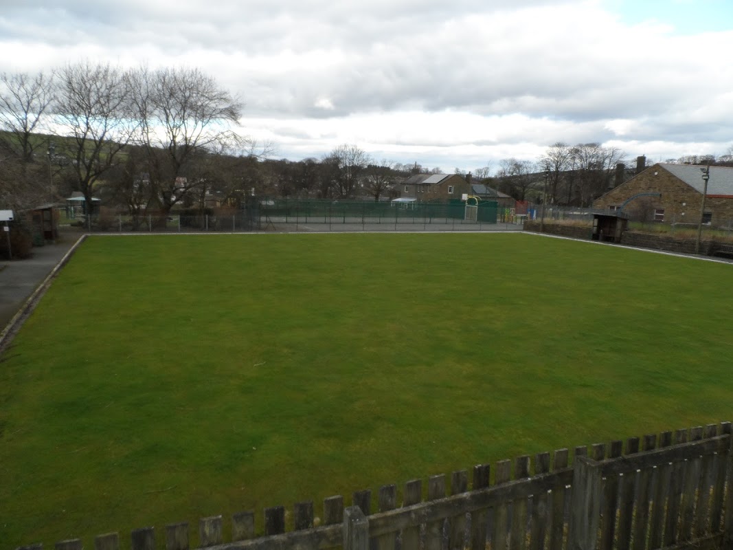 Lush green bowling green surrounded by trees and buildings under a cloudy sky in Oxenhope, Yorkshire.