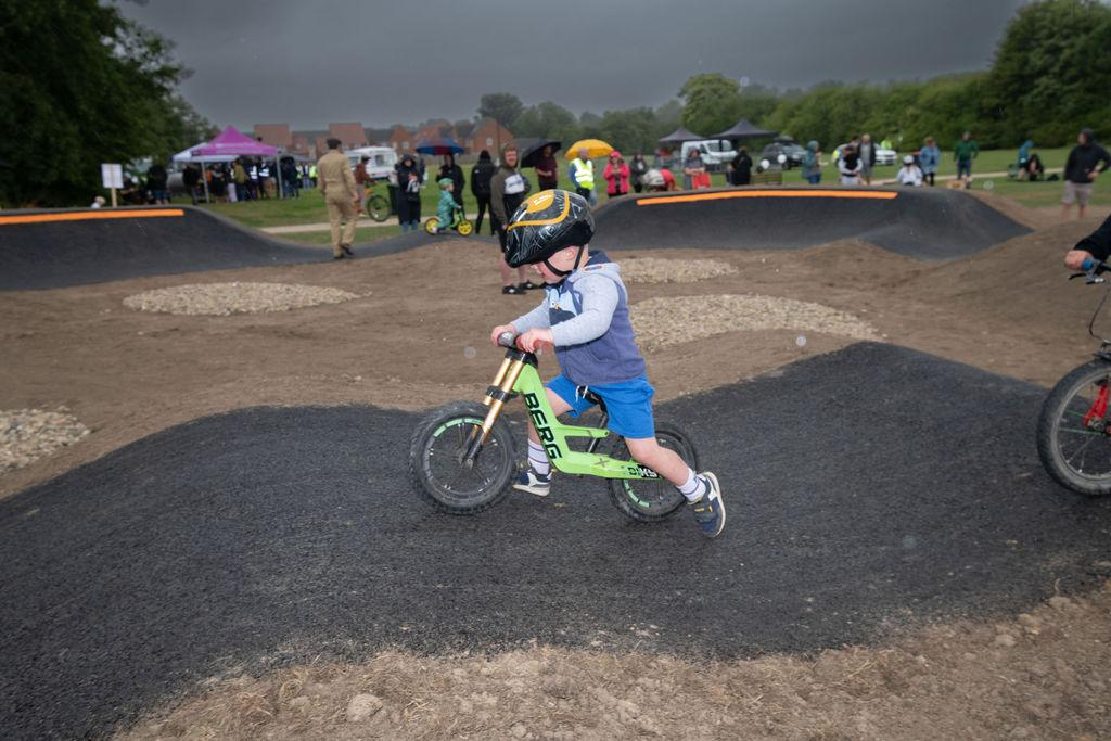 A child on a green balance bike rides over a pump track, with spectators and tents in the background.