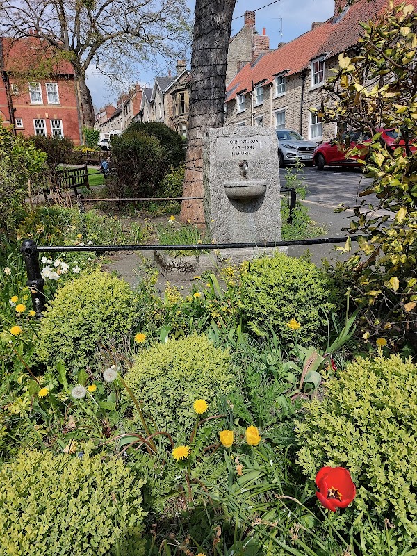 Granite memorial stone surrounded by lush greenery and colorful flowers in a quaint Yorkshire street.