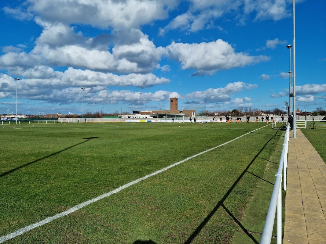 Green football pitch with white lines, a blue sky, and clouds; background features a building and a tower.