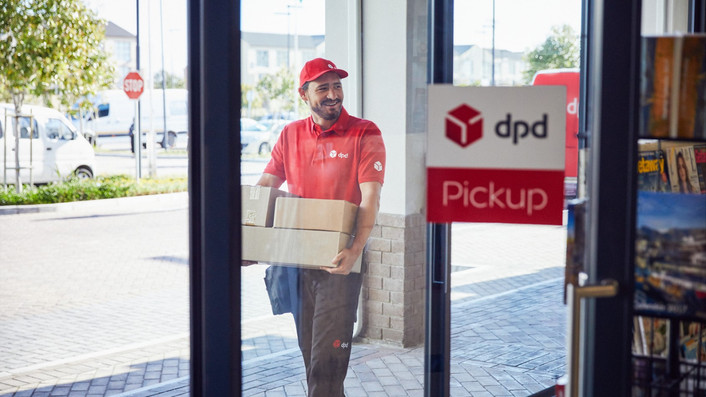 A man in a red DPD uniform carries a parcel through a glass door marked "Pickup" in a sunny parking area.