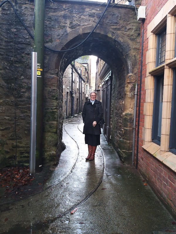 Narrow stone alleyway with a person standing under an arch, flanked by brick and stone buildings, wet pavement.