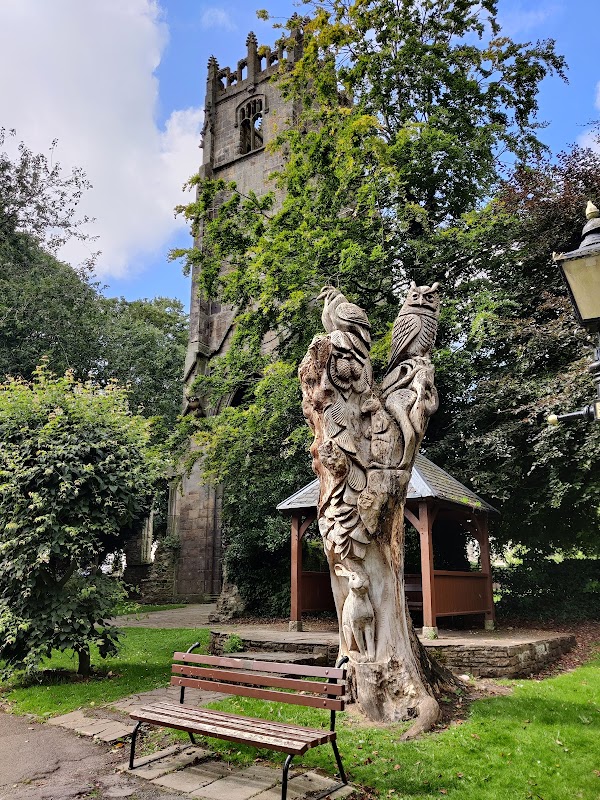 Car park with pay & display signs, surrounded by trees and a carved wooden sculpture near a church tower.