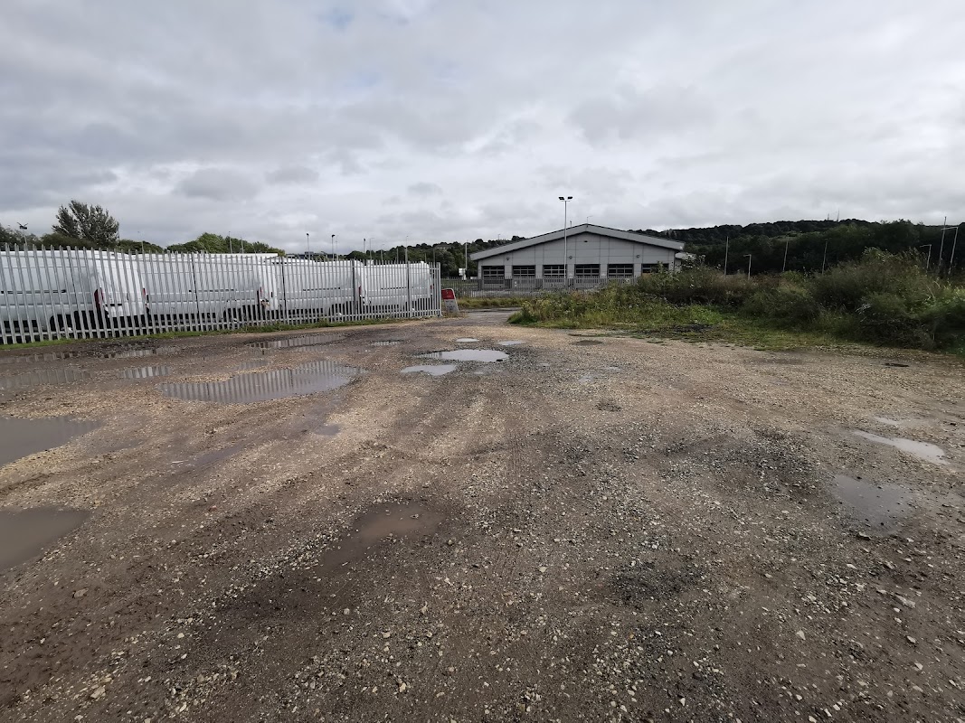 Gravel car park with puddles, surrounded by greenery and a building in the background, near Centenary Riverside Nature Park.