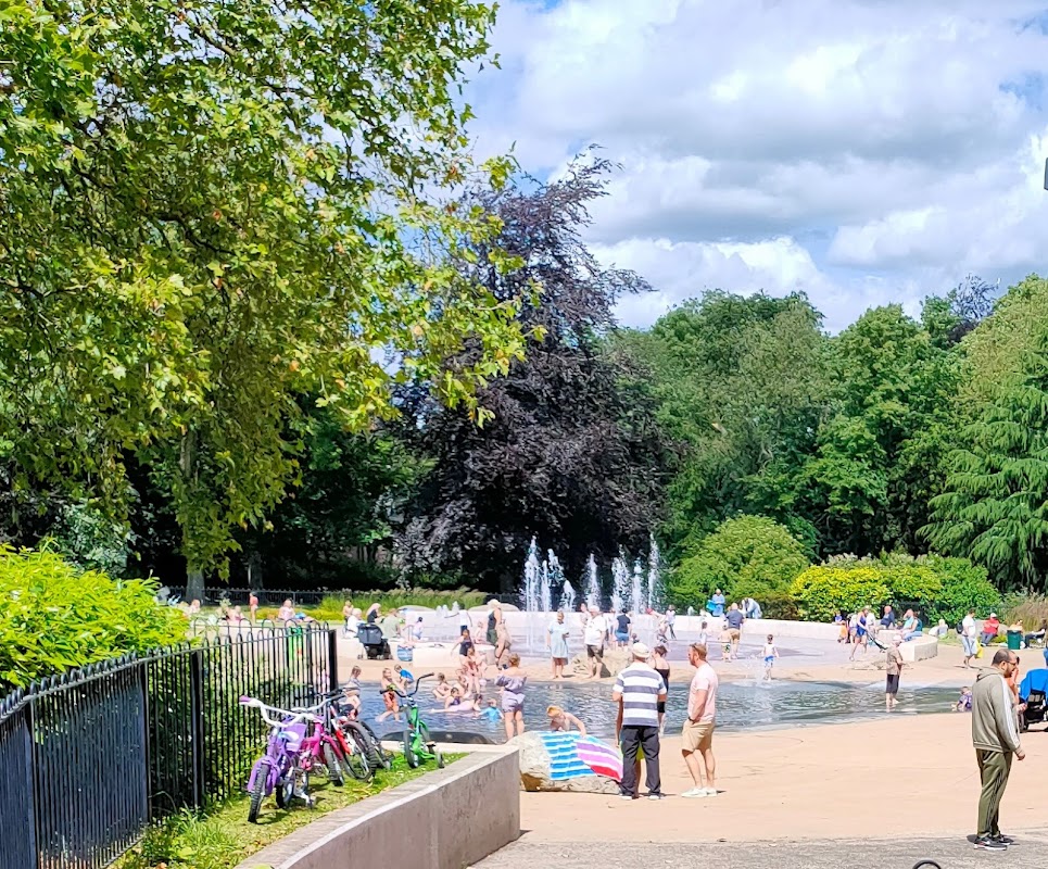 Families enjoying a sunny day at Clifton Park, with a splash pad, greenery, and bicycles nearby.