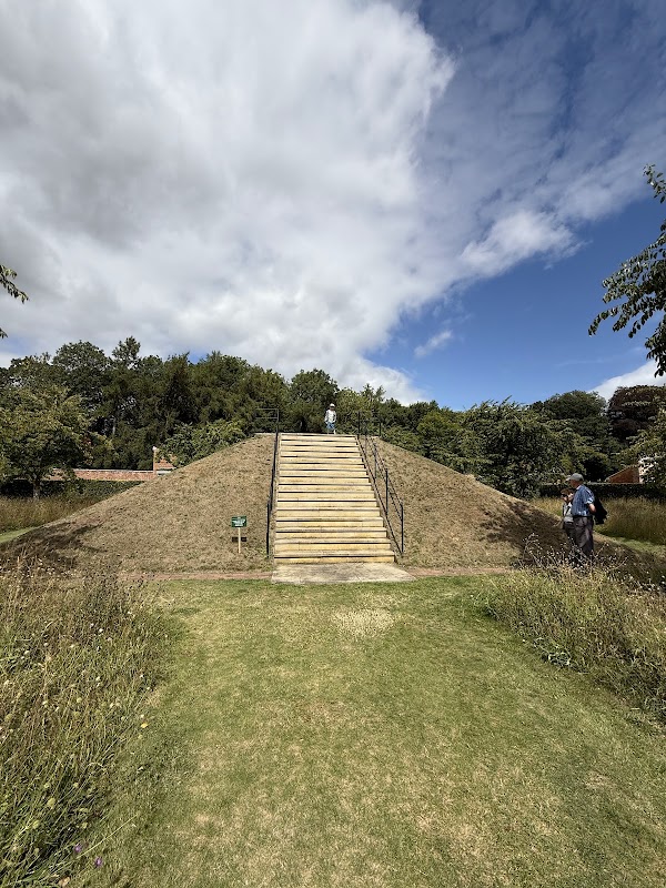 Staircase leading up a grassy mound, surrounded by lush greenery and under a partly cloudy sky.
