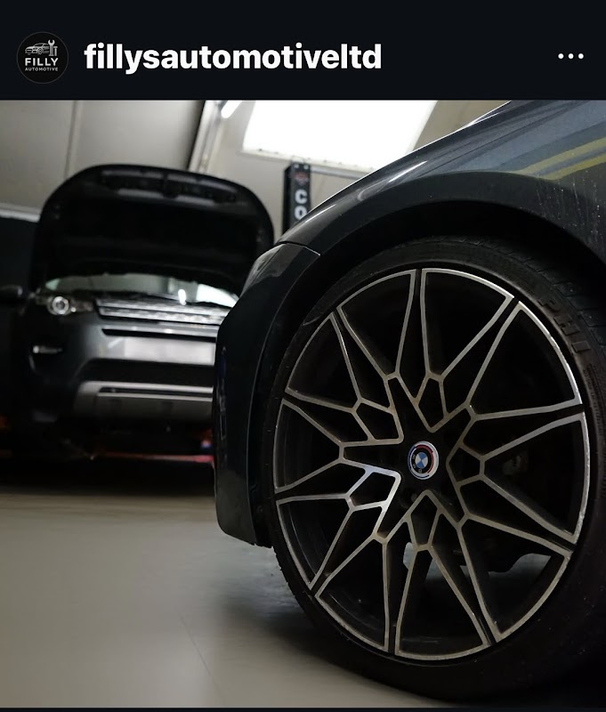 Close-up of a stylish alloy wheel with a blurred car in the background and another vehicle with an open hood.