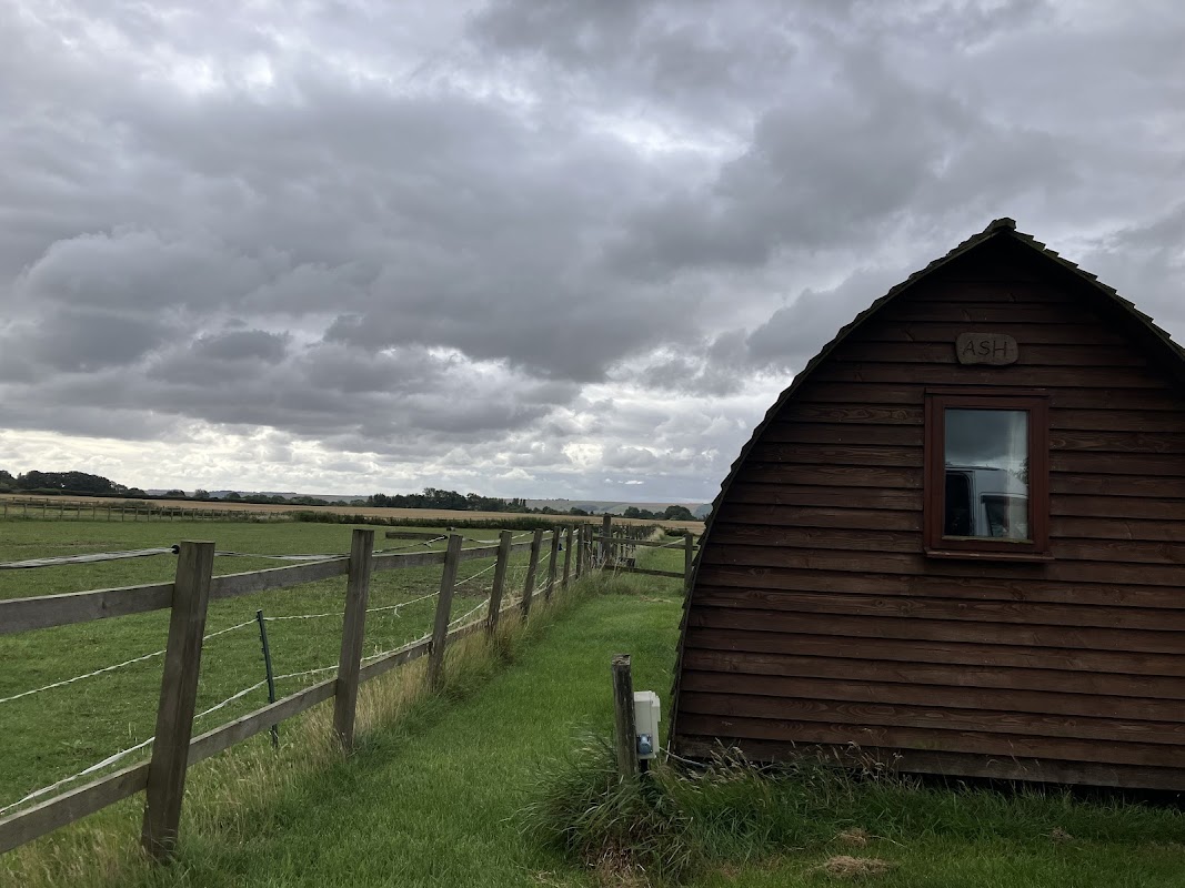 Wooden cabin named "ASH" beside a grassy field, under a cloudy sky with distant hills and a wooden fence.