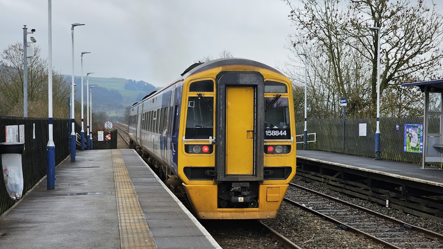 Train approaching a platform at Giggleswick station, surrounded by trees and hills in the background.