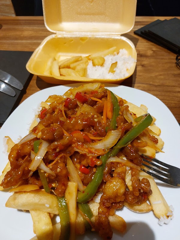 A plate of stir-fried chicken with vegetables and fries, next to a takeout container with rice and more fries.