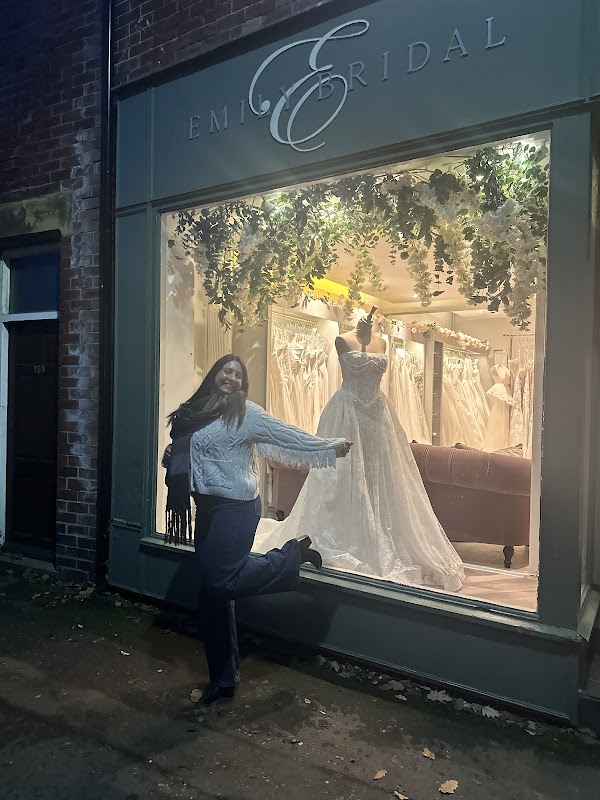 A woman poses joyfully outside a bridal shop featuring elegant wedding dresses in the window display.
