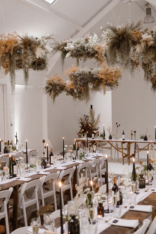 Elegant floral arrangements hang overhead in a bright venue, with long tables set for a celebration, featuring candles and wine.
