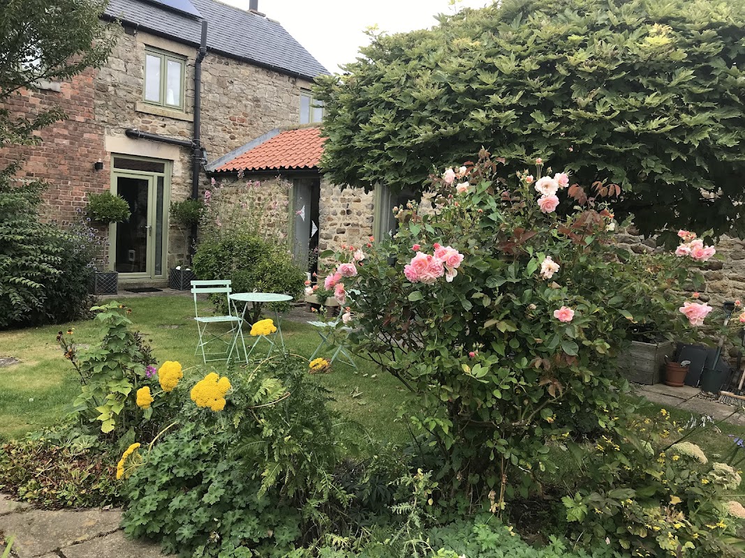 Charming guest house with stone walls, vibrant flower garden, and green seating area under a lush tree.