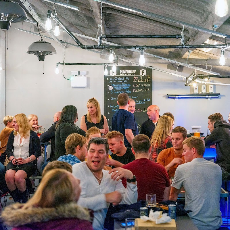 Lively brewery interior with patrons socializing, a bar, and a chalkboard menu in a modern, industrial setting.