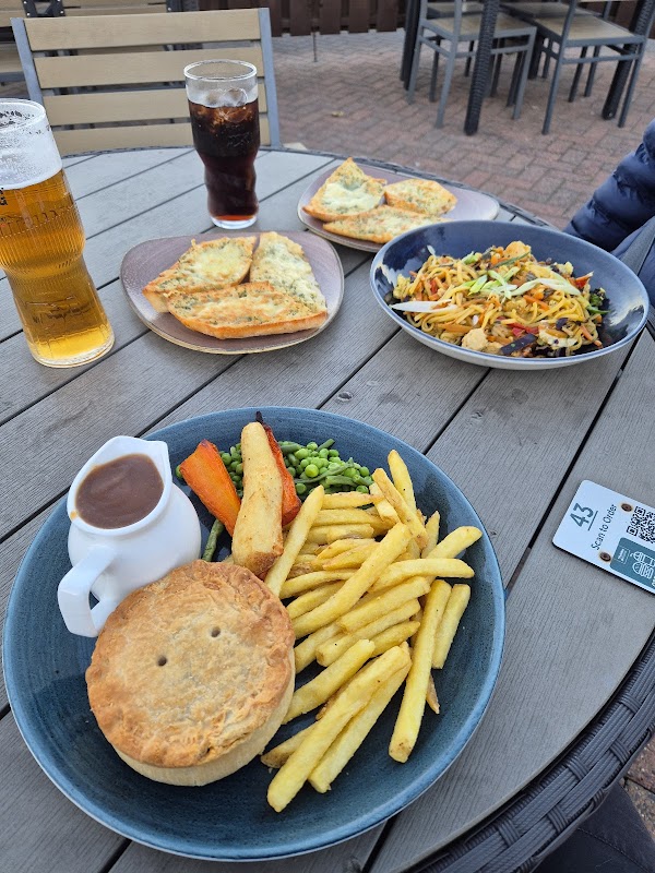 A table set with a pie, fries, peas, carrots, and salad, alongside drinks and garlic bread in a casual cafe setting.