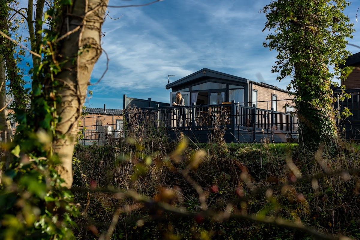 A modern lodge with a deck overlooks a tranquil setting, surrounded by trees and blue skies at Willow Pastures Country Park.