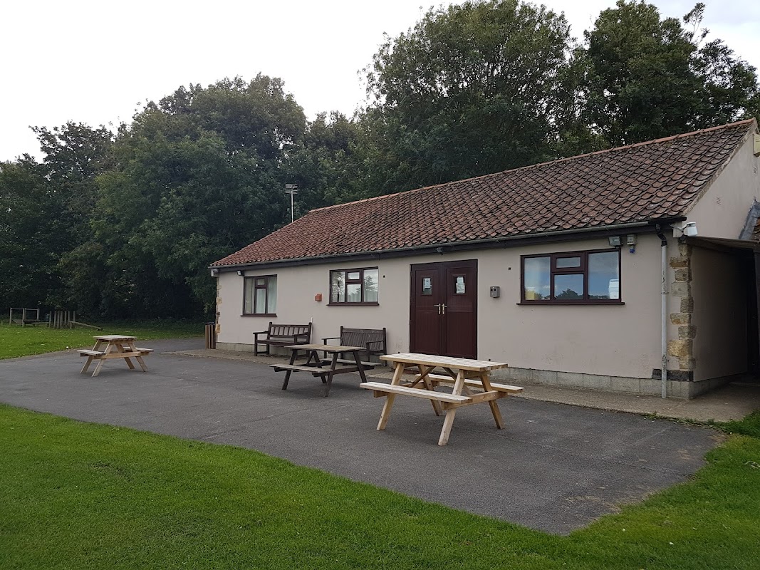 A sports club building with a brown door, picnic tables, and green grass, surrounded by trees in Slingsby, Yorkshire.