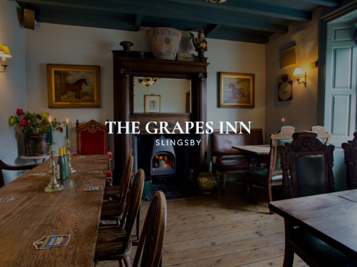 Cozy interior of The Grapes Inn with wooden tables, chairs, a fireplace, and framed artwork on the walls.