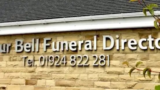 Sign for Arthur Bell Funeral Directors, featuring contact number and stone wall background.