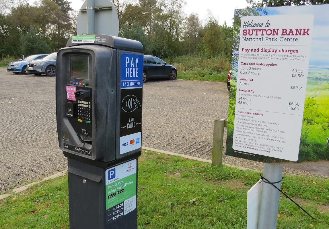 Pay & Display machine and sign detailing parking charges at Sutton Bank Car Park, surrounded by greenery and vehicles.