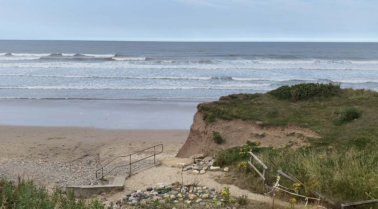 A sandy beach with gentle waves, grassy cliffs, and a rocky path leading to the shoreline under a cloudy sky.