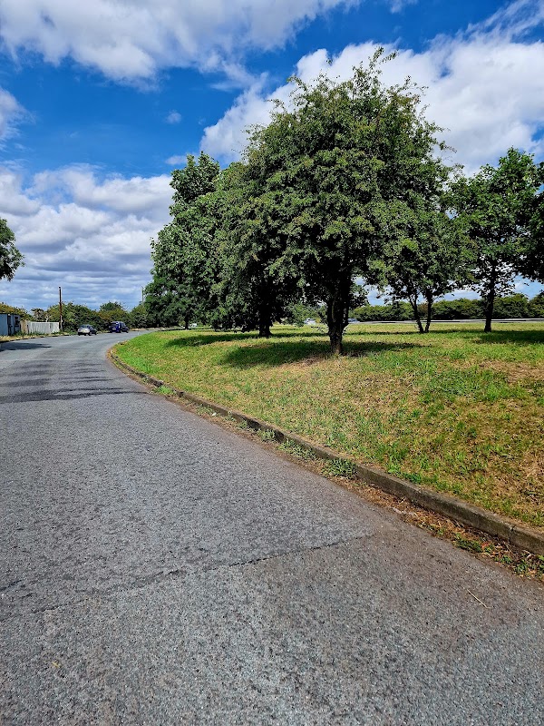 Curved road lined with trees and grassy areas, with a cloudy blue sky above in Thimbleby, North Yorkshire.