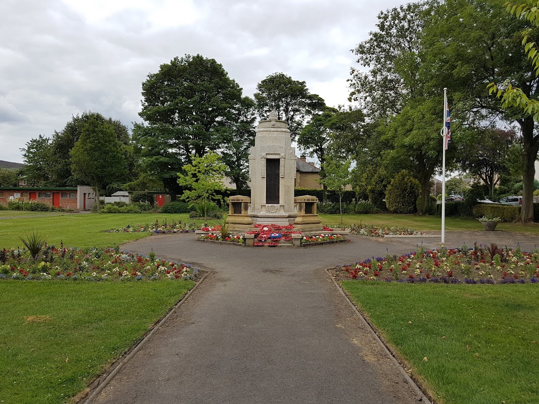 A landscaped park featuring a war memorial, flower beds, and trees under a cloudy sky.
